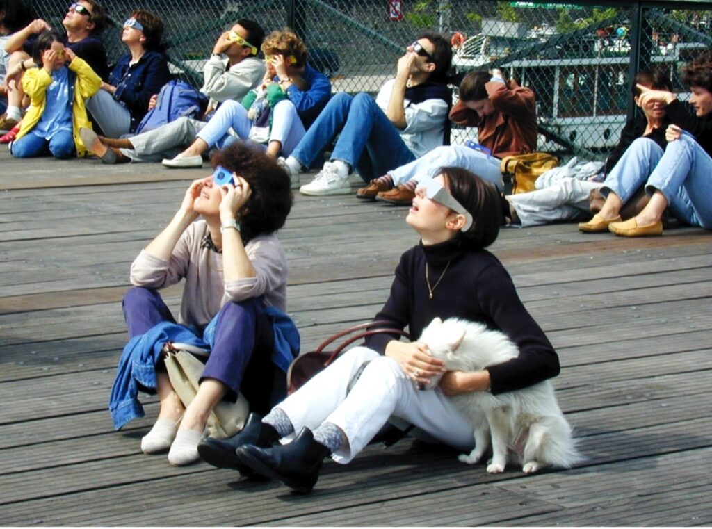 In the foreground, two women are sat on the floor looking up at the sky. They are wearing eclipse viewers, which look like thick glasses made from cardboard. One of the women is hugging a white dog and covering the dog’s eyes. Other people wearing similar eclipse viewers also sit on the floor in the background
