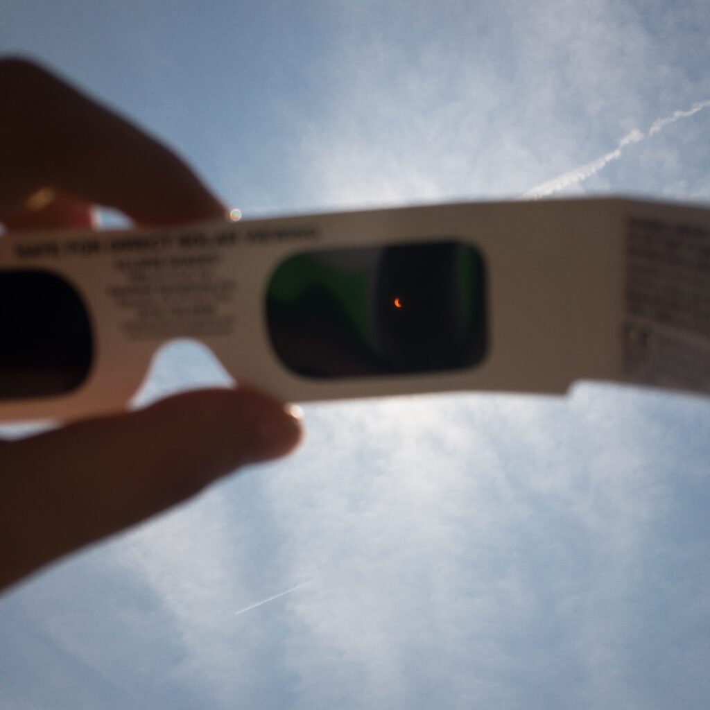 A hand holds a pair of boxy cardboard glasses in the middle of a blue sky in the background. The eye lenses are black, with one of them showing a tiny orange crescent shape.